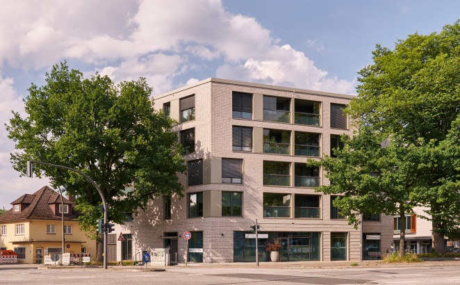 Modernes Eckgebäude mit heller Klinkerfassade und großen Fenstern an einer belebten Straßenkreuzung mit großen Bäumen.