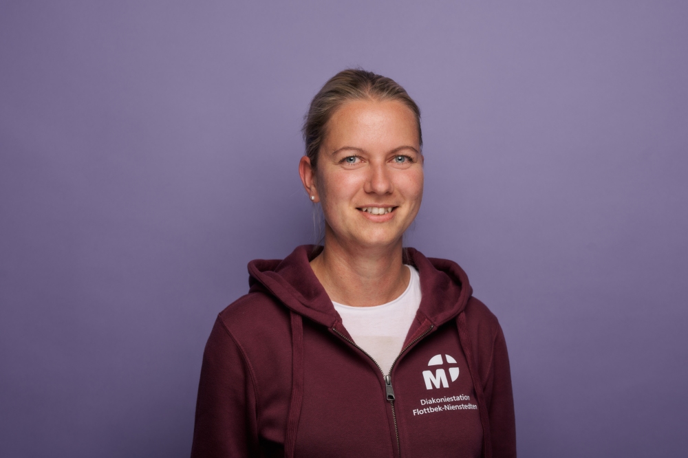Porträt einer lächelnden Frau mit blondem Haar vor einem violetten Hintergrund. Sie trägt einen dunkelroten Hoodie mit Logo der Diakoniestation Flottbek-Nienstedten.