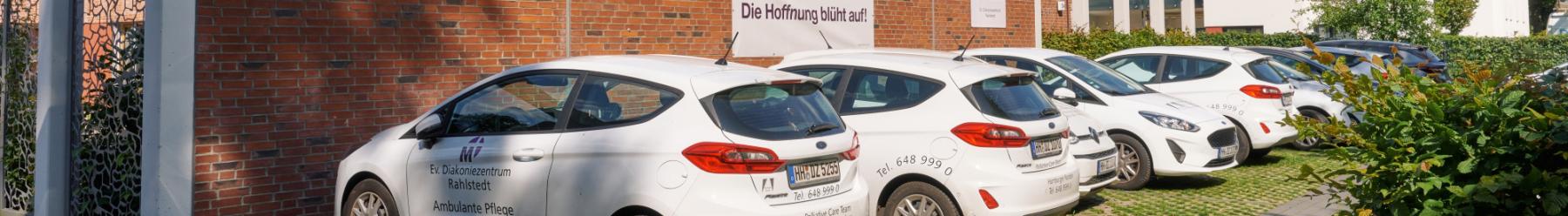 Parkplatz mit mehreren weißen Autos des Ev. Diakoniezentrums Rahlstedt vor einer Backsteinwand, im Hintergrund grüne Hecken.