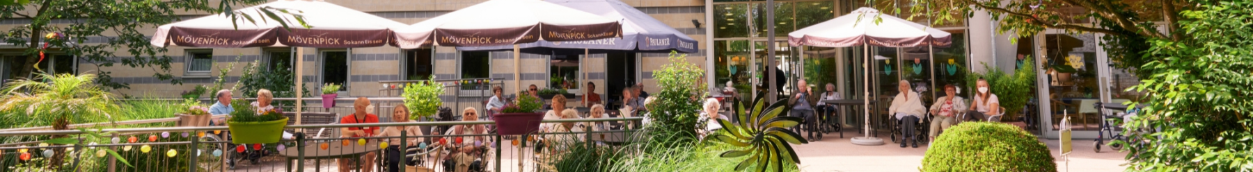 Bewohnerinnen und Bewohner genie?en einen sonnigen Tag unter Sonnenschirmen auf einer Terrasse im gr?nen Innenhof des Seniorenzentrums.