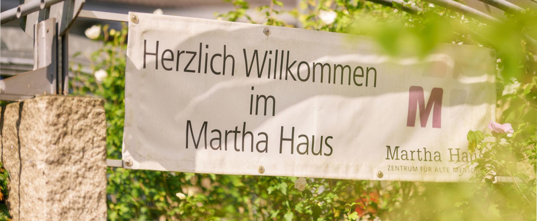 Willkommensschild mit der Aufschrift ?Herzlich Willkommen im Martha Haus?, umgeben von gr?ner Natur und sommerlichem Sonnenschein.