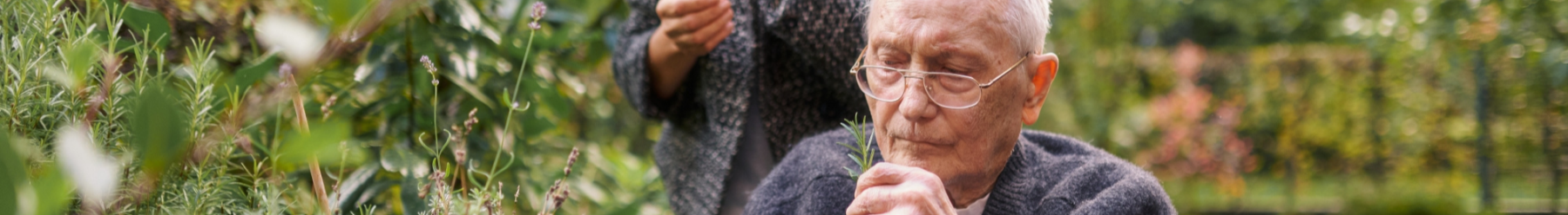 Älterer Mann im Garten riecht an einem Rosmarinzweig, im Hintergrund unscharf eine Pflegekraft und grüne Pflanzen.