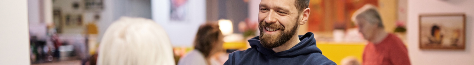 Pflegekraft mit Bart lächelt eine ältere Person an. Im Hintergrund unscharf ein Gemeinschaftsraum mit weiteren Personen.