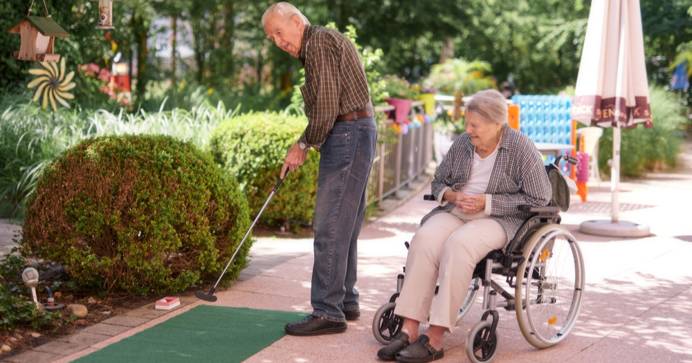 Ein älterer Mann spielt Minigolf auf einer kleinen Bahn im Garten, während eine ältere Frau im Rollstuhl ihm zuschaut.