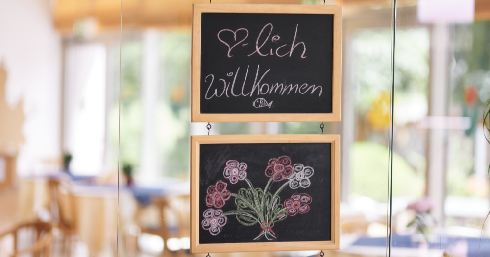 Zwei Tafeln mit handgeschriebener Begrüßung 'Herzlich Willkommen' und einer Blumenzeichnung, die an einer Glastür hängen.