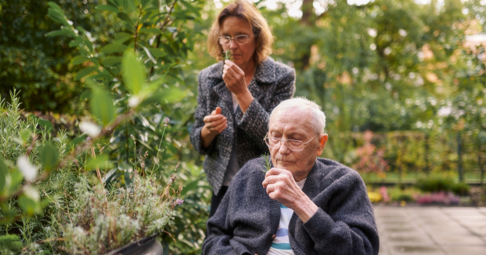 Älterer Mann im Rollstuhl riecht an einem Rosmarinzweig, Pflegekraft steht dahinter und riecht ebenfalls an einem Zweig. Beide sind umgeben von Pflanzen im Garten.