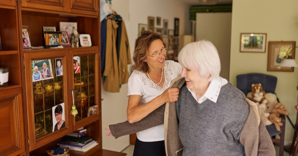 Pflegekraft hilft einer lächelnden älteren Frau beim Anziehen einer Jacke, im Hintergrund ein Schrank mit Fotos und Deko.