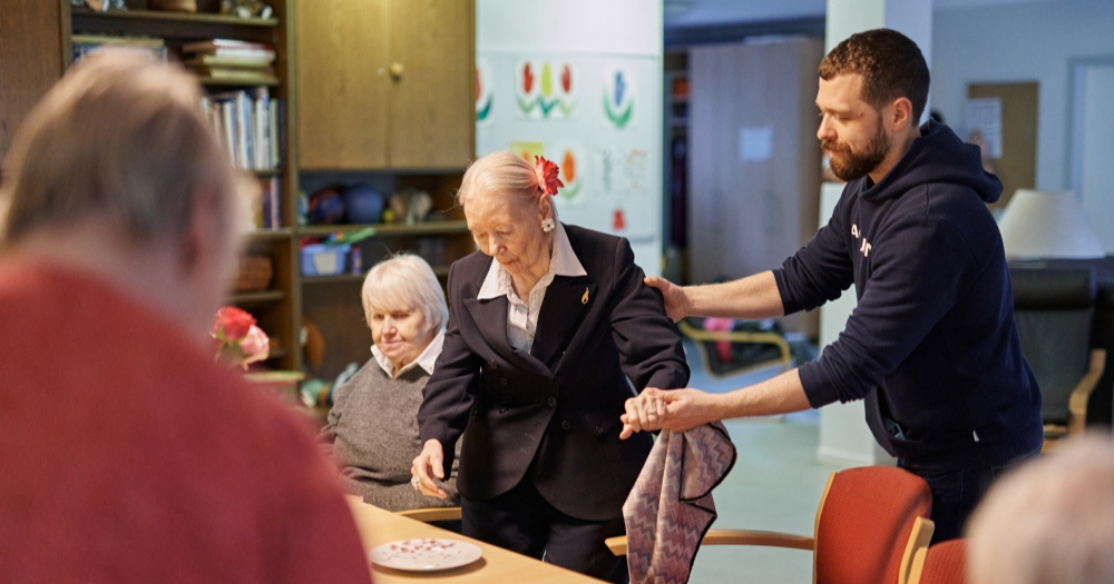 Männliche Pflegekraft unterstützt eine ältere Frau beim Hinsetzen. An dem Tisch in einem Gemeinschaftsraum sitzen weitere Personen.
