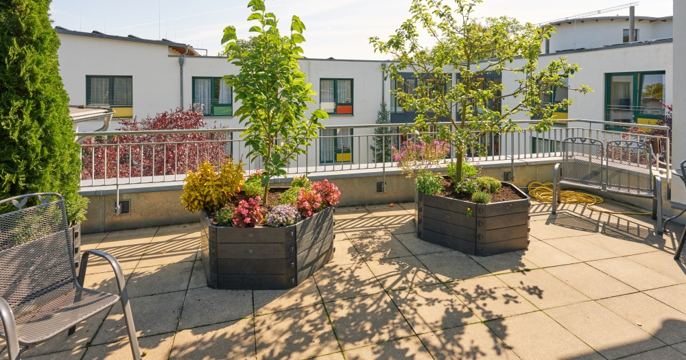 Sonnige Dachterrasse mit Blumenk?beln, B?umen und Sitzgelegenheiten. Sie ist umgeben von modernen Geb?uden mit farbigen Fenstern.