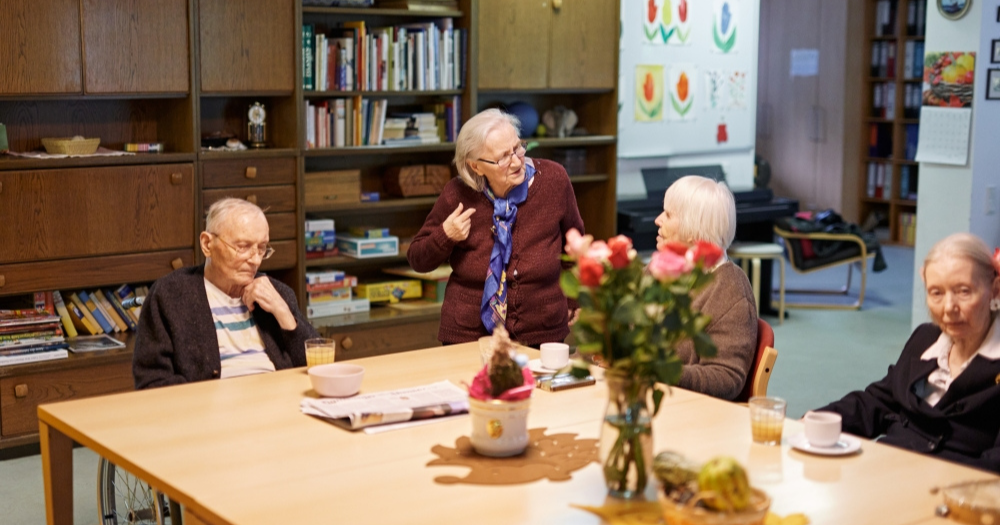 Ältere Menschen sitzen an einem Tisch in einem Gemeinschaftsraum. Eine Frau steht und spricht. Im Hintergrund ein Wohnzimmerschrank aus dunklem Holz mit Bücherregal.