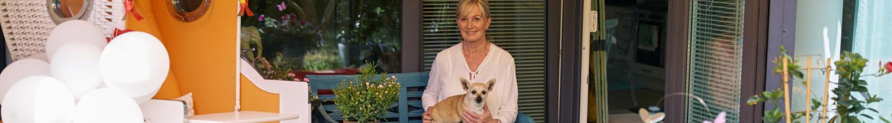Eine Frau sitzt mit einem Hund auf dem Scho? auf einer Terasse mit Strandkorb sowie Dekorationen und Pflanzen in leuchtenden Farben.
