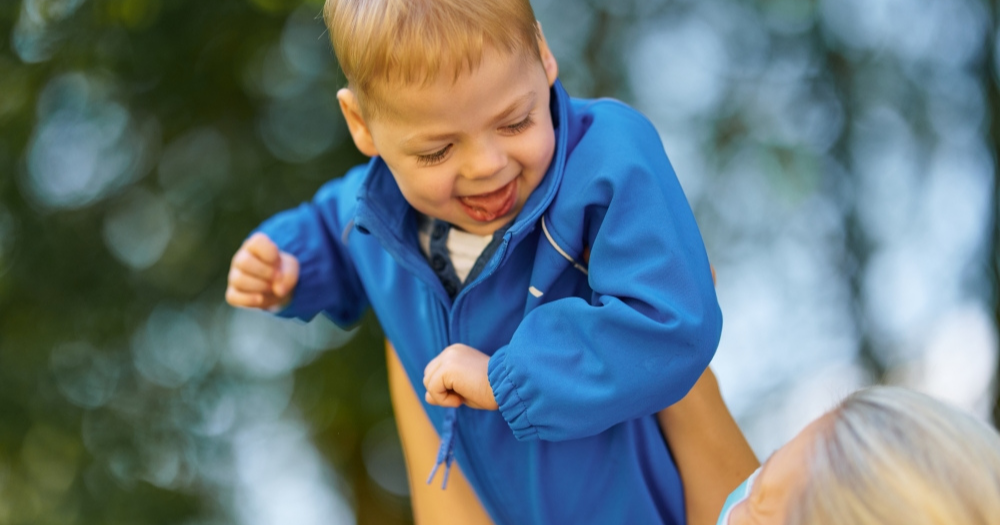 Ein lachender Junge in blauer Jacke wird von einer Person hochgehoben. Das Bild erzählt von Freude, Geborgenheit und spielerischer Interaktion im Freien.