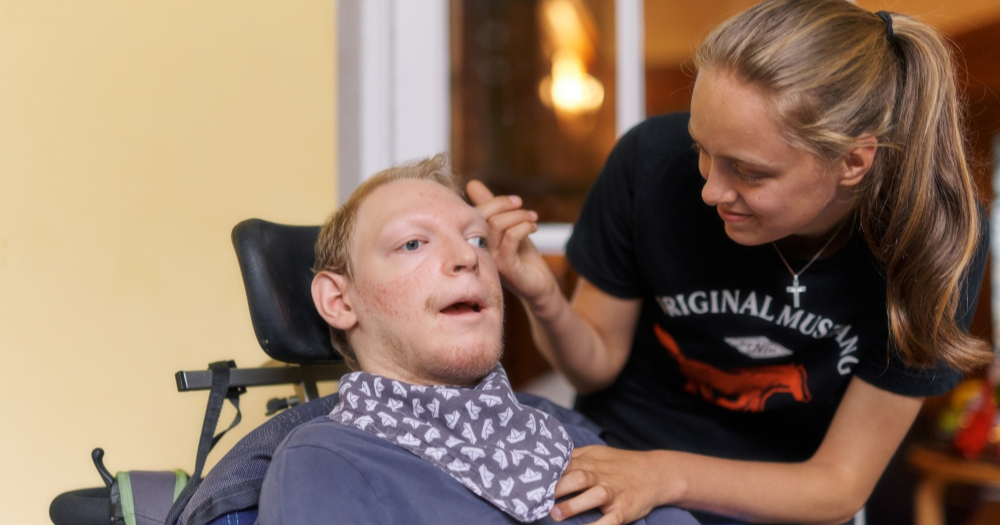 Junger Mann im Rollstuhl mit Halstuch, betreut von einer jungen Frau mit Pferdemotiv-Shirt. Sie blickt ihn an und hält behutsam ihre Hand an seine Wange.
