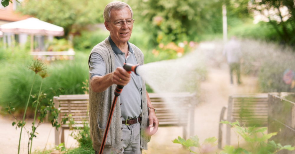 Älterer Mann mit grauem Haar gießt mit einem Gartenschlauch Pflanzen in einer grünen Gartenanlage. Er trägt eine Weste und lächelt vergnügt.